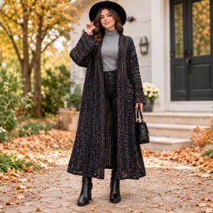 May include: A long, open-front black crochet cardigan with a textured, loose-knit pattern. The outfit includes a grey mock-neck top, black high-waisted jeans, a black wide-brimmed hat, black leather ankle boots, and a small black handbag.