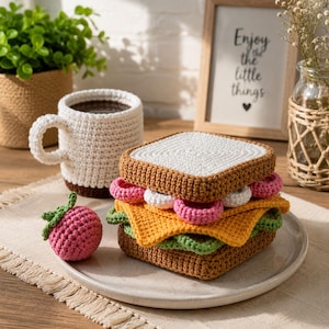 Puede incluir: Un juguete de sándwich de ganchillo con rebanadas de pan marrón, una capa de queso amarillo, lechuga verde, rodajas de tomate rosa y aros de cebolla blancos, acompañado de una pequeña fruta de ganchillo rosa y una taza blanca tejida.