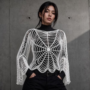 May include: A white crochet long-sleeve top featuring a prominent spiderweb pattern that radiates from the center of the chest. The garment has wide, flared sleeves and a scalloped hemline, worn over a solid black long-sleeve shirt and black pants.