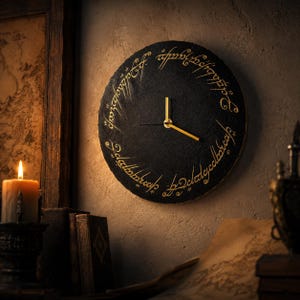 Puede incluir: Un reloj de pared redondo negro con manecillas doradas y una inscripción en una fuente estilizada. El reloj está montado en una pared texturizada, con una vela encendida y libros en el fondo. El diseño del reloj evoca un tema de fantasía.