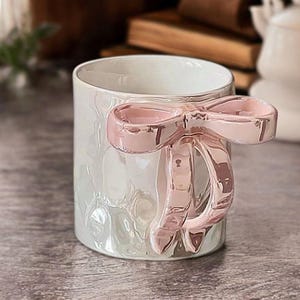 May include: A white ceramic mug with a pearlescent, hammered texture finish. The mug features a prominent, three-dimensional handle shaped like a large, glossy, metallic light pink bow that extends from the side of the cylindrical vessel.