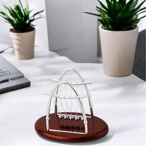 May include: A Newton's cradle featuring five polished silver metal spheres suspended by thin lines from two arched silver frames, all mounted on a smooth, dark brown oval wooden base, creating a classic physics demonstration device.