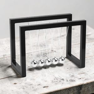 Peut inclure: Un pendule de Newton composé de cinq sphères métalliques argentées polies, suspendues par de fins fils blancs à un cadre rectangulaire noir. L'objet présente deux montants noirs parallèles reliés au sommet, maintenant les billes en équilibre.