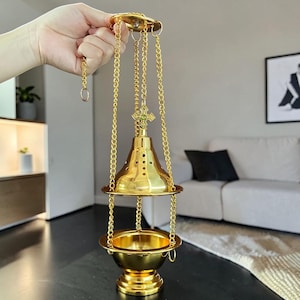 Könnte beinhalten: Ein hängendes Weihrauchfass aus goldfarbenem Metall mit glockenförmigem Deckel, der Perforationen zur Belüftung aufweist. Es besitzt ein kleines Kreuzornament und eine schalenförmige Basis, gehalten von drei Ketten an einem Ring.