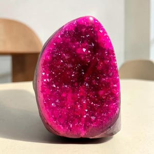 Op de afbeelding: Een levendig felroze geode-exemplaar met een cluster van kleine, sprankelende kristallen in een uitgeholde steenholte. De buitenkant van de steen is glad en donker, wat contrasteert met de intense, verzadigde magentakleur van de binnenkant.