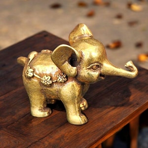 May include: A small, metallic gold-colored elephant figurine with a textured surface. The elephant features large ears, a raised trunk, and a decorative band across its back adorned with a series of small, raised flower-shaped embellishments.