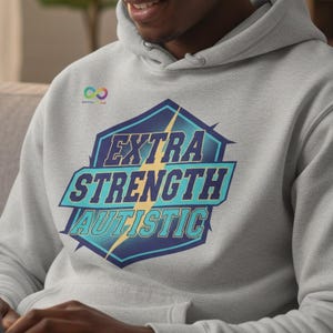 May include: A light gray hoodie with a graphic that reads "EXTRA STRENGTH AUTISTIC" in a blue and teal design. The hoodie has a drawstring hood and a small logo on the upper left chest.