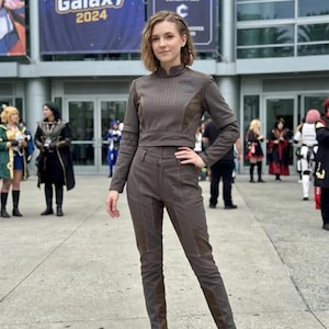 Op de afbeelding: Een persoon in een bruin cosplay-outfit, bestaande uit een top met lange mouwen en een broek. De outfit heeft donkerbruine accenten en wordt gecombineerd met bruine laarzen. Op de achtergrond staat een banner met "Cosplay Galaxy 2024".