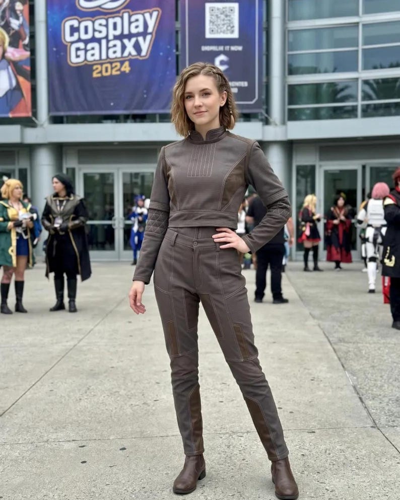 Op de afbeelding: Een persoon in een bruin cosplay-outfit, bestaande uit een top met lange mouwen en een broek. De outfit heeft donkerbruine accenten en wordt gecombineerd met bruine laarzen. Op de achtergrond staat een banner met "Cosplay Galaxy 2024".
