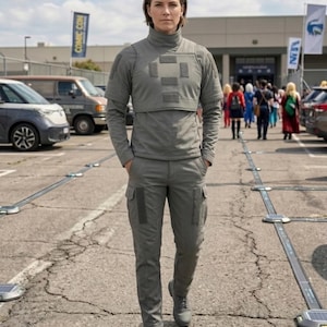 May include: A person wearing a gray tactical outfit, including a high-neck top with a chest panel and cargo pants. The outfit is a solid gray color, and the person is standing in a parking lot.