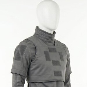 Puede incluir: Un chaleco t&aacute;ctico gris con cuello alto y mangas cortas se muestra en un maniqu&iacute;. El chaleco presenta m&uacute;ltiples parches cuadrados y costuras reforzadas. El dise&ntilde;o sugiere un enfoque en la protecci&oacute;n y la funcionalidad.