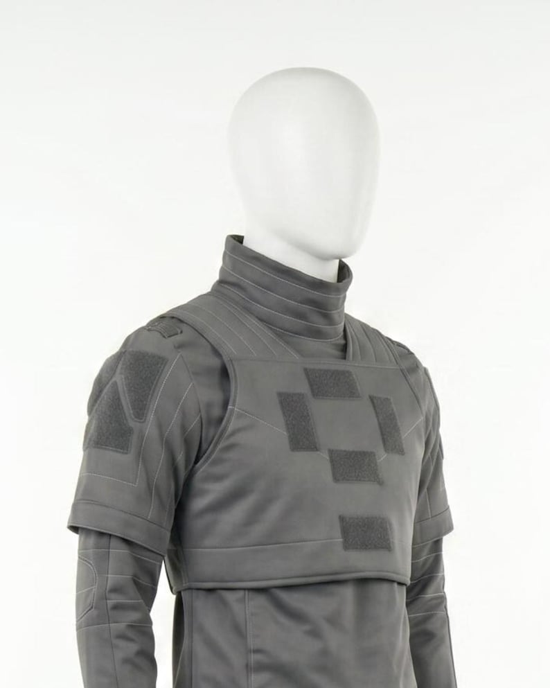 Puede incluir: Un chaleco t&aacute;ctico gris con cuello alto y mangas cortas se muestra en un maniqu&iacute;. El chaleco presenta m&uacute;ltiples parches cuadrados y costuras reforzadas. El dise&ntilde;o sugiere un enfoque en la protecci&oacute;n y la funcionalidad.