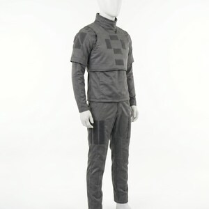 Puede incluir: Un uniforme t&aacute;ctico gris exhibido en un maniqu&iacute;. El atuendo incluye una parte superior de cuello alto con una placa en el pecho, mangas largas y pantalones a juego. El uniforme tiene parches cuadrados y &aacute;reas reforzadas.