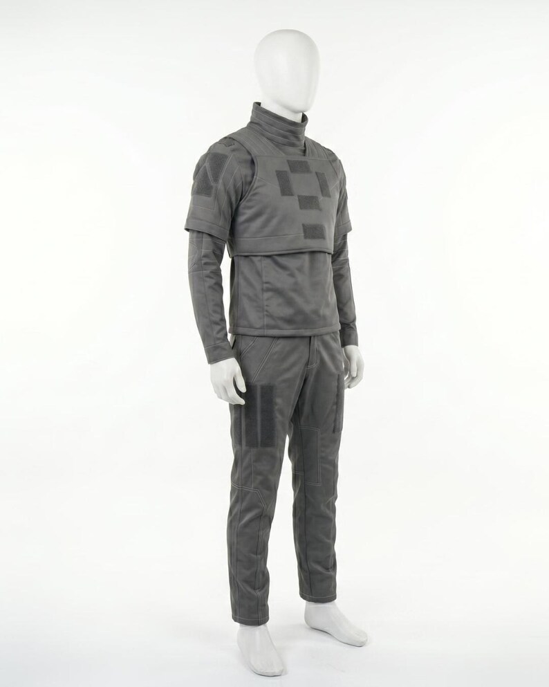 Puede incluir: Un uniforme t&aacute;ctico gris exhibido en un maniqu&iacute;. El atuendo incluye una parte superior de cuello alto con una placa en el pecho, mangas largas y pantalones a juego. El uniforme tiene parches cuadrados y &aacute;reas reforzadas.