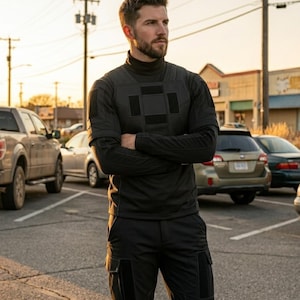 K&ouml;nnte beinhalten: Eine Person tr&auml;gt ein schwarzes Langarmshirt mit hohem Kragen und eine schwarze Cargohose. Das Shirt hat quadratische Patches auf Brust und Schultern. Die Person steht auf einem Parkplatz mit Autos im Hintergrund.