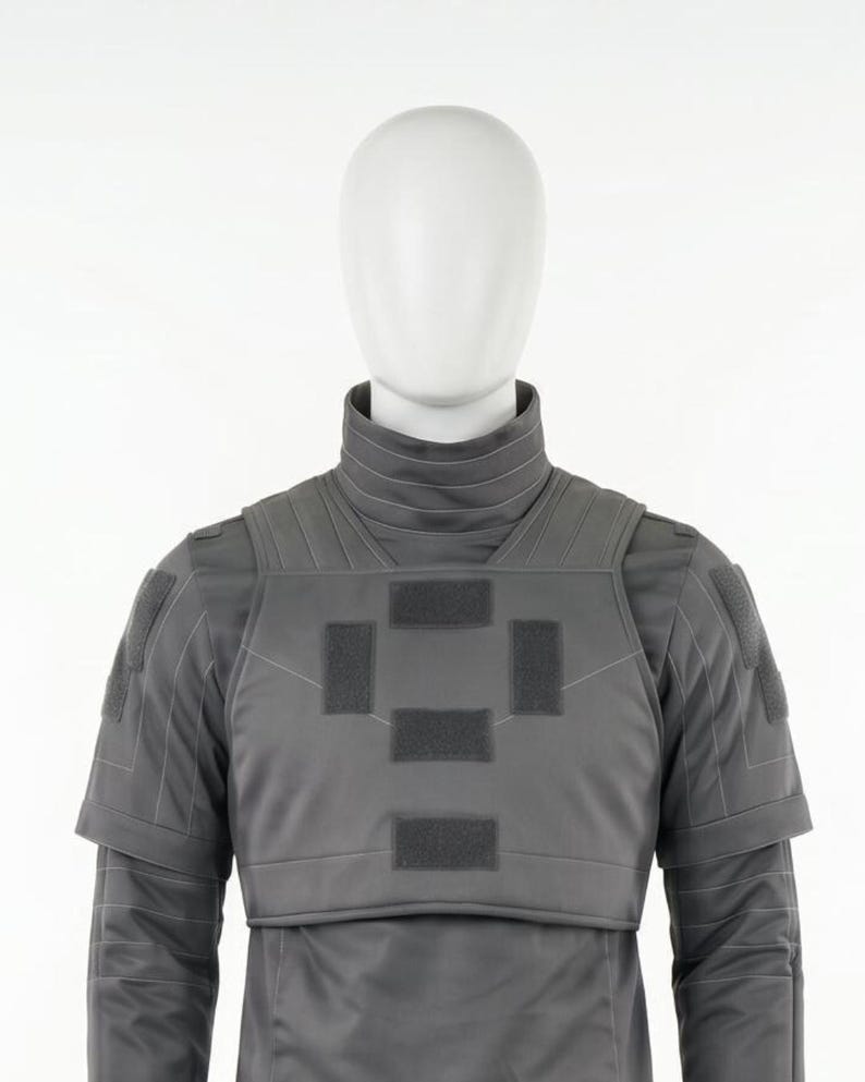 Puede incluir: Un chaleco t&aacute;ctico gris con cuello alto y mangas cortas se muestra en un maniqu&iacute;. El chaleco presenta m&uacute;ltiples parches rectangulares y est&aacute; hecho de un material liso y mate. La cabeza del maniqu&iacute; es blanca.