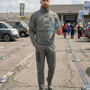 Puede incluir: Una persona con un atuendo t&aacute;ctico gris, que incluye una parte superior de cuello alto, pantalones y zapatos. La persona sostiene una tableta transparente con una interfaz futurista. El fondo muestra un estacionamiento con autos y personas, y una pancarta de Comic Con.
