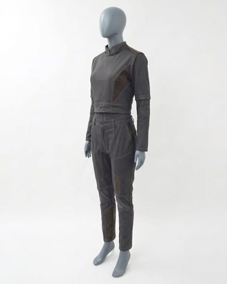 Op de afbeelding: Een donkergrijze tweedelige outfit op een mannequin. De set bestaat uit een top met hoge hals en lange mouwen en een bijpassende broek. De outfit heeft bruine accenten op de schouders, ellebogen en knie&euml;n. De broek heeft een getailleerd ontwerp.