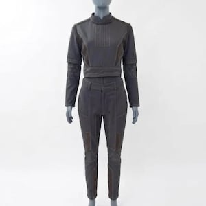 Op de afbeelding: Een tweedelige outfit in donkergrijs, getoond op een mannequin. De top is een cropped jasje met lange mouwen en een hoge kraag. De broek is getailleerd met patches op de knie&euml;n. De outfit heeft zichtbare stikseldetails.