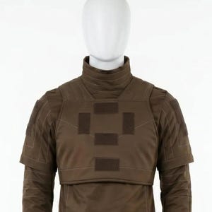 May include: A brown tactical vest with a high collar and shoulder pads, displayed on a mannequin. The vest features multiple rectangular patches and a layered design. The long-sleeved garment is a solid brown color, suitable for military or tactical applications.