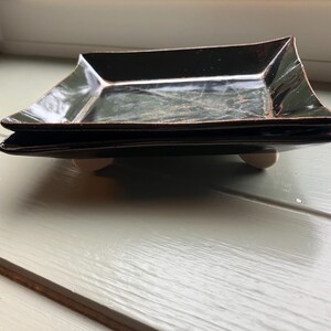 May include: A square, dark brown ceramic tray with a glossy finish and slightly raised edges. The tray is supported by four small, light-colored feet at the corners, elevating the base slightly above the surface it rests upon.