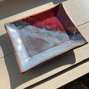 May include: A handmade ceramic square dish with a glossy finish. The dish features a gradient of colors, transitioning from a deep red center to a vibrant blue rim. Decorative leaf-like patterns are embossed along the edges.