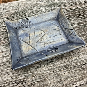 May include: A rectangular ceramic dish with a blue and gray leaf design. The dish has a raised rim and is decorated with embossed leaf patterns and textures. The color palette includes shades of blue, gray, and hints of gold.