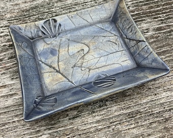 May include: A rectangular ceramic dish with a blue and gray leaf design. The dish has a raised rim and is decorated with embossed leaf patterns and textures. The color palette includes shades of blue, gray, and hints of gold.