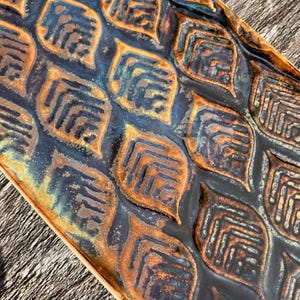 May include: A rectangular ceramic tray featuring a repeating embossed leaf pattern. The surface displays a gradient of deep navy blue and metallic bronze tones, with the raised leaf edges highlighted in a warm, burnished copper finish.