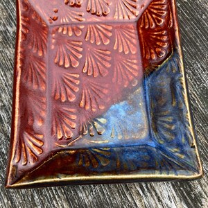 May include: Square ceramic dish with a raised, repeating fan-like pattern. The dish is primarily a deep reddish-brown color, transitioning to a blue hue on one corner. The edges are slightly rounded and have a golden-brown trim.