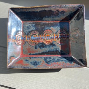 May include: Rectangular ceramic dish with a glossy finish. The dish features a blend of blue, red, and brown hues. The center has a series of embossed circular designs. The corners are slightly rounded. The dish is likely handmade.