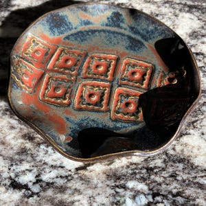 May include: A handmade ceramic plate with a unique design. The plate features a combination of colors, including blue, orange, and gold. The plate has a square pattern in the center and a wavy edge. The plate is approximately 8 inches in diameter.