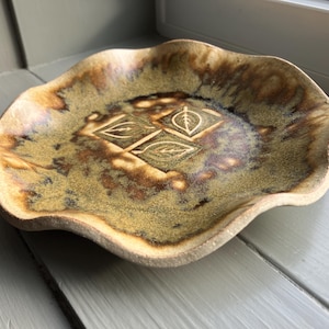 May include: A handmade ceramic dish with a wavy rim. The dish features a leaf design in the center and is glazed in shades of brown, green, and tan. The dish is approximately 8 inches in diameter.