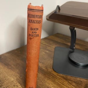 Może przedstawiać: Pomarańczowa książka zatytułowana "Elementary Analysis" autorstwa Dakin i Porter. Grzbiet książki zawiera tytuł i nazwiska autorów w czarnym tekście. Książka stoi pionowo na drewnianej powierzchni. Nazwa wydawcy, Bell, znajduje się na dole.