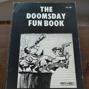 Peut inclure: Un livre vintage intitulé "THE DOOMSDAY FUN BOOK" avec une couverture noire et des illustrations. La couverture présente un dessin animé de deux hommes, l'un avec un télescope et l'autre tenant un papier intitulé "DATA". Le livre est édité par Edward Goldsmith avec des dessins de Richard Willson.