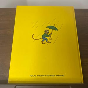 Puede incluir: Una cubierta de libro amarilla con un mono de dibujos animados sosteniendo un paraguas verde bajo la lluvia. El mono lleva un sombrero marr&oacute;n y pantalones azules. El texto "VERLAG FRIEDRICH OETINGER HAMBURG" est&aacute; impreso en la parte inferior.