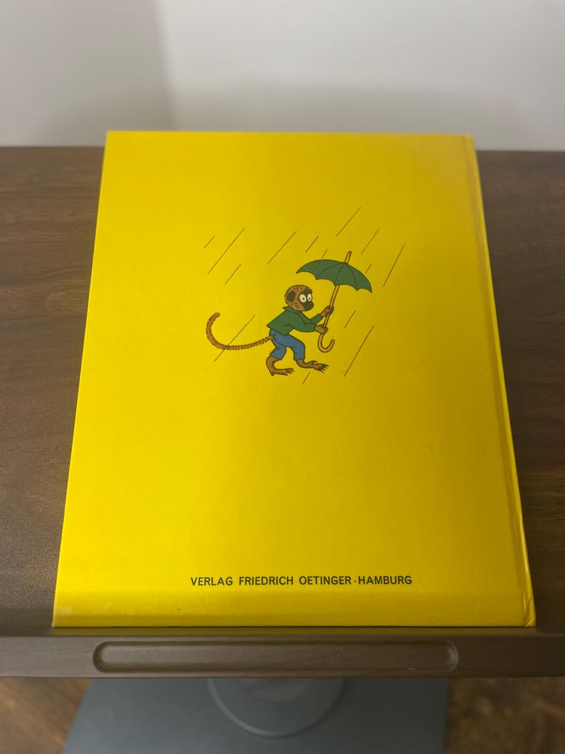 Puede incluir: Una cubierta de libro amarilla con un mono de dibujos animados sosteniendo un paraguas verde bajo la lluvia. El mono lleva un sombrero marr&oacute;n y pantalones azules. El texto "VERLAG FRIEDRICH OETINGER HAMBURG" est&aacute; impreso en la parte inferior.