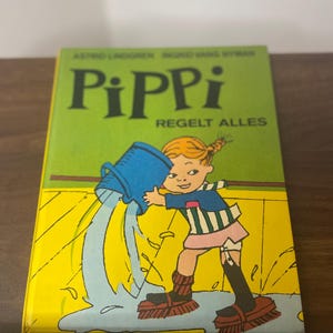Puede incluir: Un libro infantil titulado "Pippi Regelt Alles" con una portada verde. La portada presenta una ilustraci&oacute;n de una ni&ntilde;a vertiendo agua de un cubo azul. La ni&ntilde;a lleva una camisa a rayas, una falda rosa y botas negras.