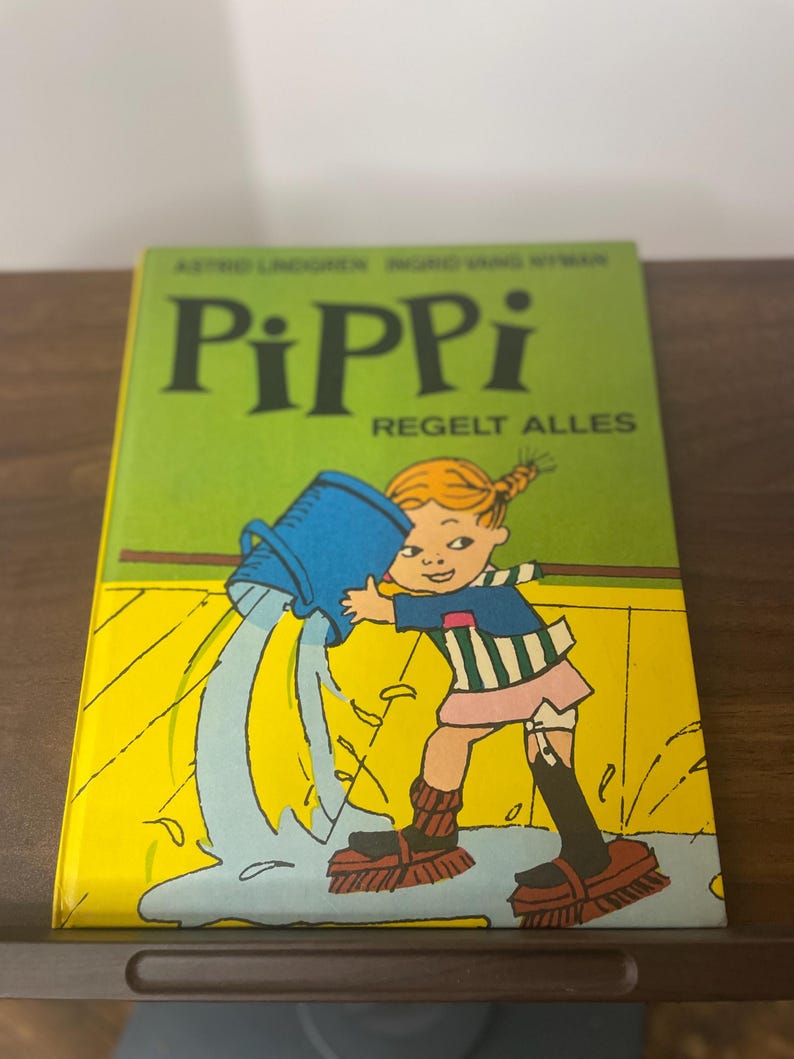 Puede incluir: Un libro infantil titulado "Pippi Regelt Alles" con una portada verde. La portada presenta una ilustraci&oacute;n de una ni&ntilde;a vertiendo agua de un cubo azul. La ni&ntilde;a lleva una camisa a rayas, una falda rosa y botas negras.