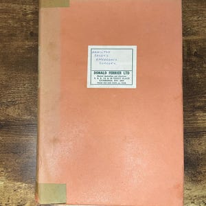 May include: A vintage book with a salmon-pink cover. The title "HAMILTON BAILEY'S EMERGENCY SURGERY" is printed on a white label. The book's spine and corners are reinforced with tan tape. The book is on a wooden surface.