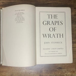 Puede incluir: Libro abierto con el título "The Grapes of Wrath" de John Steinbeck. La portada blanca tiene texto negro. La página izquierda enumera otras obras de Steinbeck. El libro está sobre una superficie de madera.