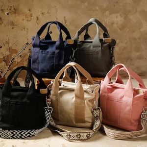 May include: Five canvas tote bags in black, navy blue, grey, beige, and pink. Each bag has a top handle and a detachable patterned shoulder strap. The bags are displayed on a wooden surface against a neutral background.
