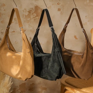 Pode incluir: Três bolsas de ombro de couro nas cores bege, preto e castanho penduradas em ganchos de metal. Cada bolsa tem uma alça longa com fivela e fecho de zíper. As bolsas são exibidas contra uma parede texturizada esbranquiçada.