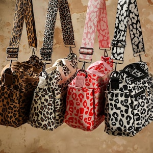 以下が含まれることがあります： 茶色、黒と白、赤のヒョウ柄のバッグが4つ。各バッグには、お揃いの柄のストラップが付いています。バッグはニュートラルな背景に吊り下げられています。バッグには複数のポケットとジッパーが付いています。