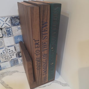 May include: A wooden cookbook holder with three cookbooks. The cookbooks are titled "Thyme to Eat", "The Taming of the Stew", and "Limited Copy Edition". The holder is made of dark wood and sits on a white countertop.