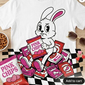 May include: White t-shirt featuring a cartoon rabbit surrounded by bags of pink chips. The rabbit is holding a bag of chips with the text "PINK CHIPS". The shirt has a black and white checkered pattern at the bottom.