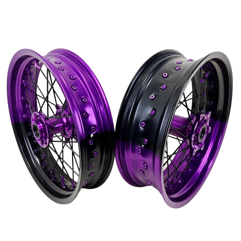 May include: Two motorcycle rims with a gradient finish, transitioning from black to vibrant purple. The rims feature black spokes and purple hubs, showcasing a modern design. The rims are set against a white background.