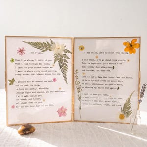 May include: A double-hinged gold metal frame containing two glass panels with pressed flowers and ferns. The left panel features the poem The Promise, and the right panel features the text I Did Think, Let's Go About This Slowly, both printed on white paper.