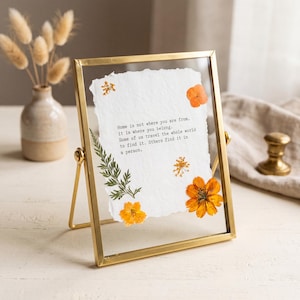 Może przedstawiać: Złota metalowa ramka typu floating frame zawierająca biały papier z postrzępionymi krawędziami. Na papierze znajduje się czarny nadruk: Home is not where you are from, it is where you belong. Some of us travel the whole world to find it. Others find it in a person. Na papierze umieszczono prasowane pomarańczowe kwiaty oraz zielony liść paproci.