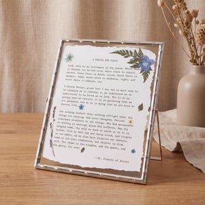 May include: A silver-toned metal frame with a bamboo-style texture containing a piece of white paper with deckled edges. The paper features the printed text of A Prayer for Peace by St. Francis of Assisi, decorated with pressed green ferns and small blue flowers.